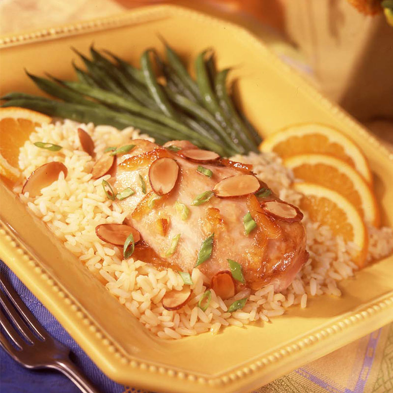 Close up view of orange chicken and rice topped with sliced almonds on a yellow plate with orange wedges and asparagus.