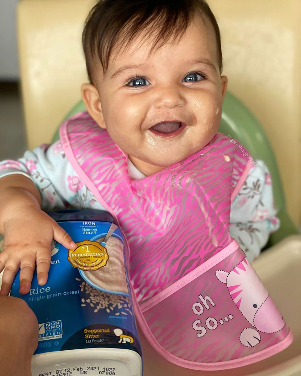 Smiling baby in high chair wearing pink bib with hand on package of infant rice cereal