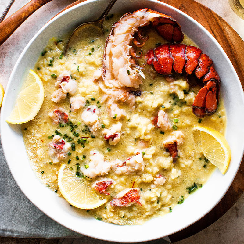 Butter Poached Lobster Risotto Overhead shot of a white bowl full of Butter Poached Lobster Risotto with exposed red lobster tails and lemon wedges.