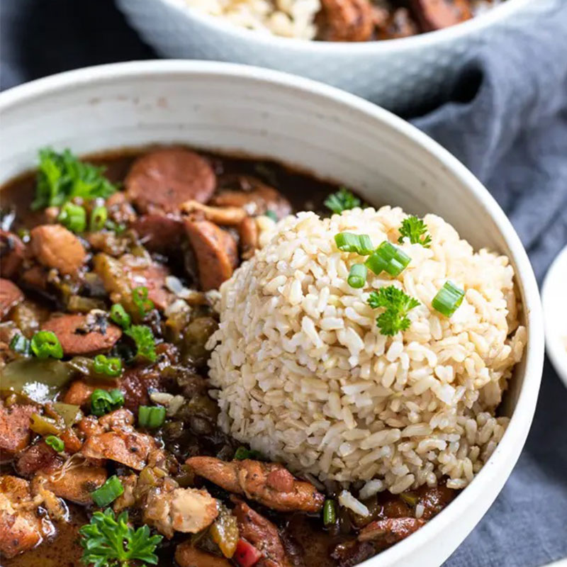 Side view of chicken and sausage gumbo with brown rice in a white bowl.