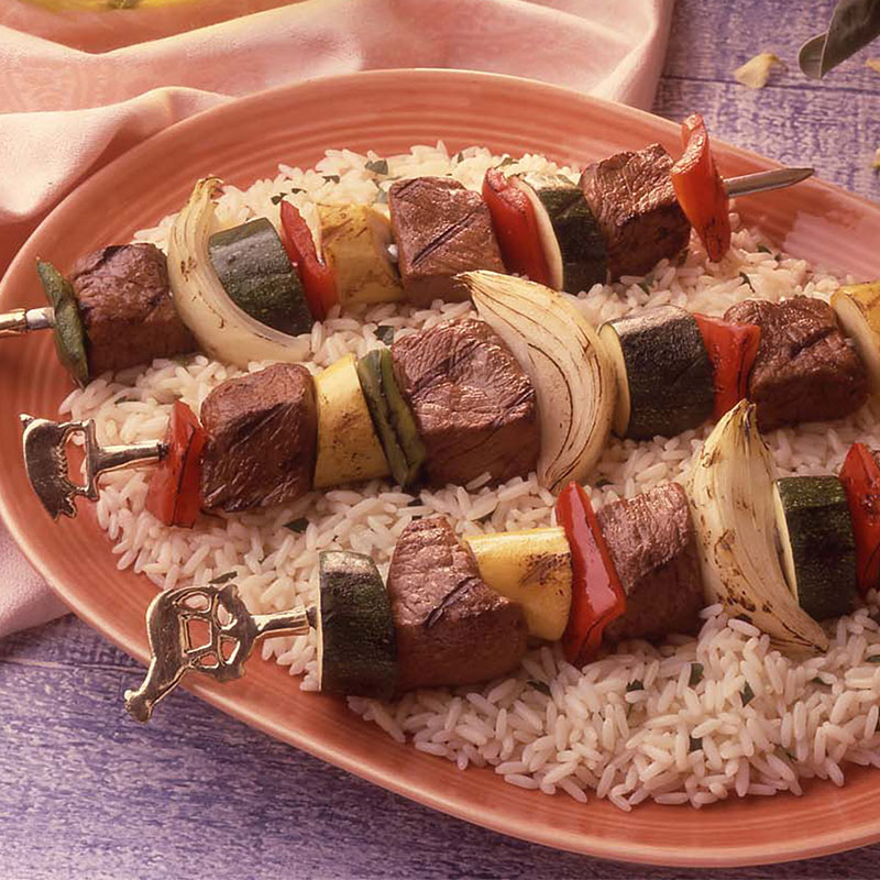 Side view of grilled beef kabobs over rice on a terracotta plate.