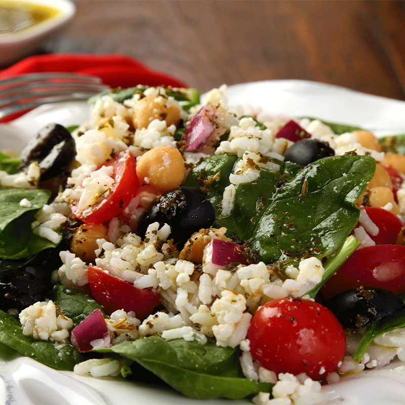 A closeup image of Mediterranean Rice Salad on a white plate with dressing in the distance.