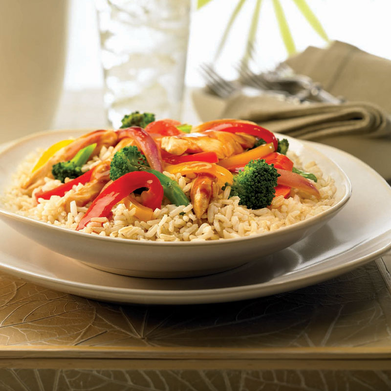 Orange Chicken and Vegetable Rice Bowl Side view of a bowl of Orange Chicken and Vegetable Rice with a glass and silverware in the distance.