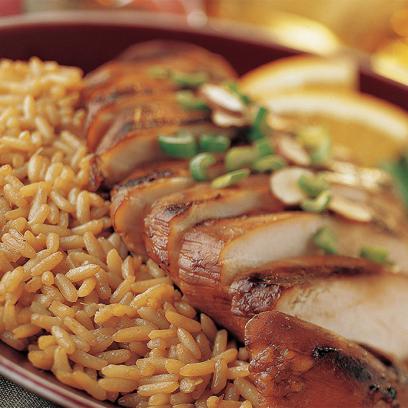 Spicy Chinese Chicken and Rice Slices of chicken sit on a bed of yellow colored rice with vegetables out of focus to the side.