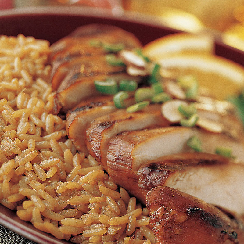 Slices of chicken sit on a bed of yellow colored rice with vegetables out of focus to the side.