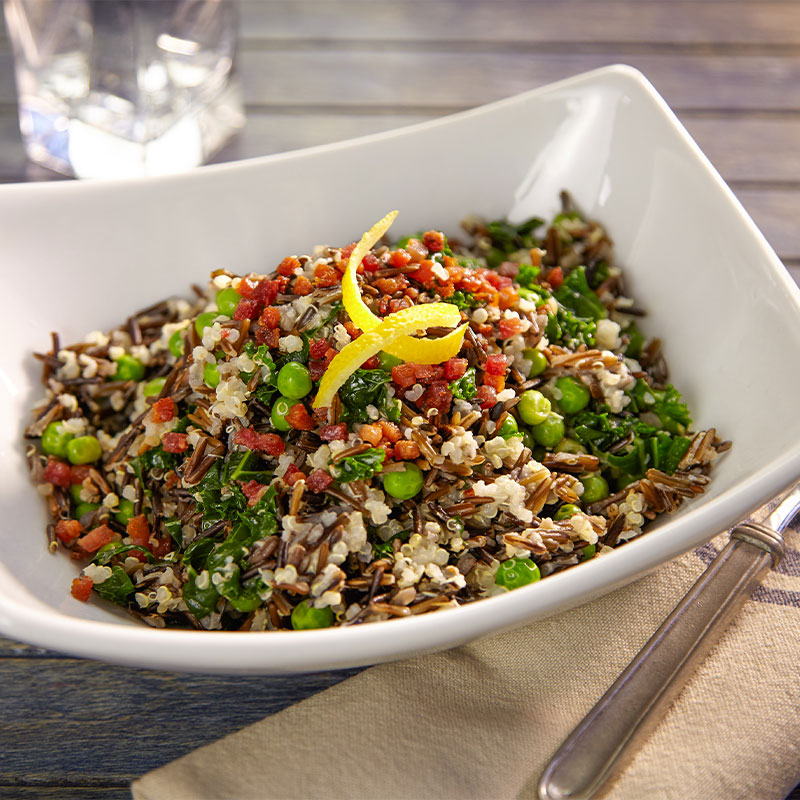 Overhead view of Spring Pilaf with Pancetta in a large white serving bowl topped with a lemon peel slice.