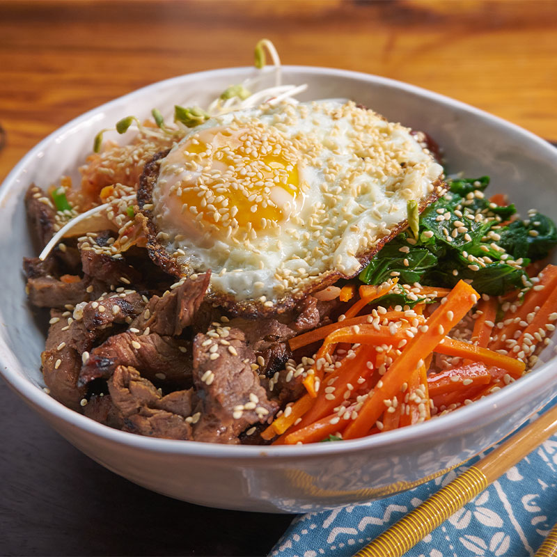 Overhead look at a colorful Steak Bibimbap with a fried egg on top in a white bowl.