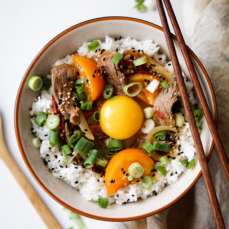 Overhead shot of a bowl with a bed of white rice topped with stir fried orange tomatoes, beef, and an egg yolk. 