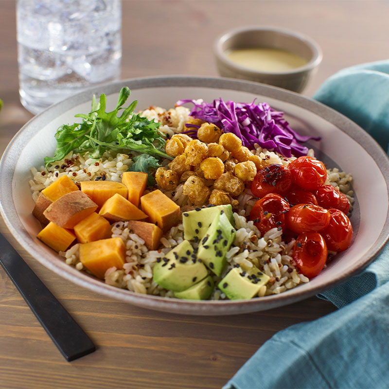 A brown rice bowl topped with chickpeas, avocado pieces, red cabbage, sweet potato chunks, and baby red tomatoes.