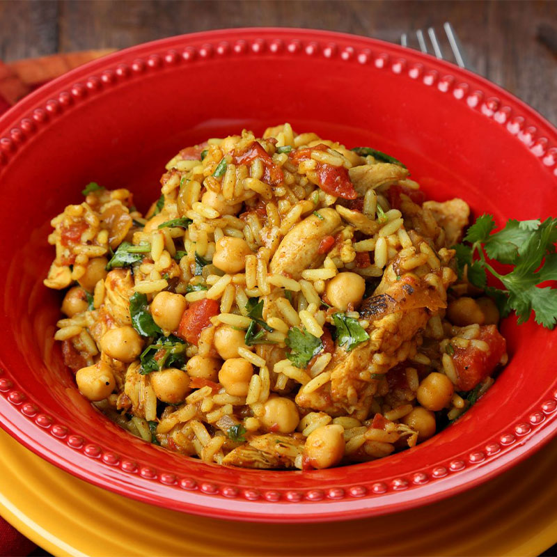 Super Quick Moroccan Chicken and Rice Overhead shot of yellow chicken and rice in a red bowl with chickpeas and parsley on top.