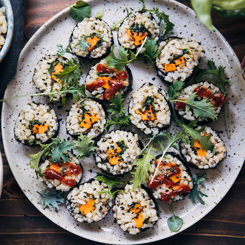 Sweet Potato Sushi Overhead shot of 15 pieces of Sweet Potato Sushi on a plate with sushi topped with garnish of microgreens.