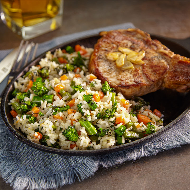 A close up image of toasted garlic rice with broccoli and carrot pieces exposed next to a porch chop on a skillet.