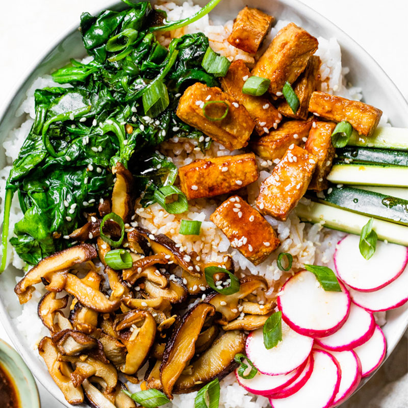 Overhead image of white rice topped with spinach, cooked tofu, mushrooms, and cucumbers in a white bowl.