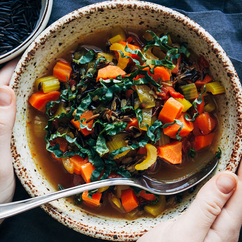 Overhead view of a hand holding a bowl full of Wild Rice Soup with big pieces of carrot and celery exposed. 