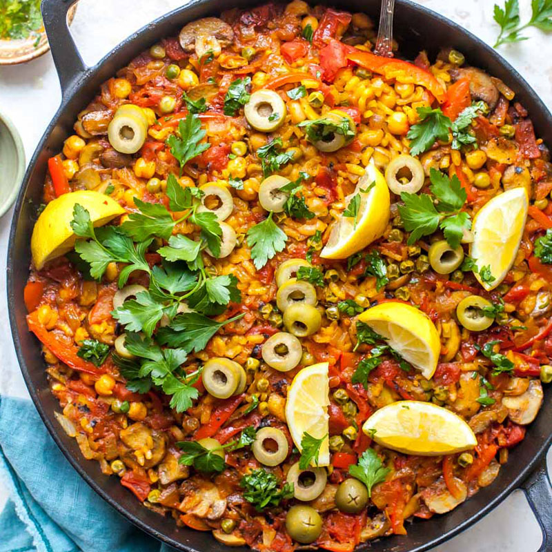 Vegetarian Paella Overhead shot of a skilled filled with colorful rice paella with parsley, lemons, and olives visible on top.