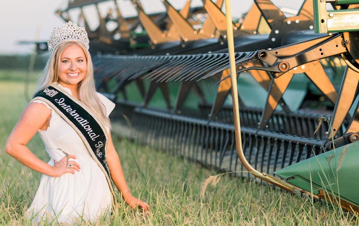 84th-IRF-Queen-Caroline-Hardy standing in mature rice field next to green combine