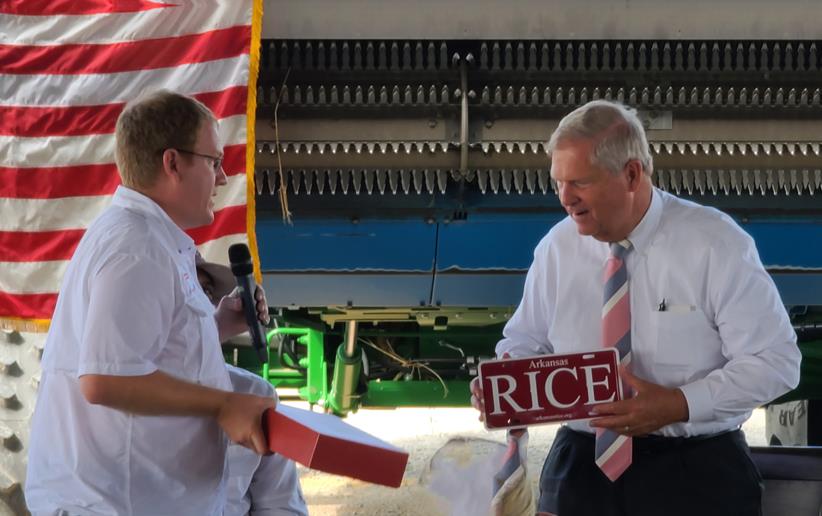 Two men stand in fron of American flag and combine, the one on the right, USDA Secretary Tom Vilsack, holds a R-I-C-E signholds an