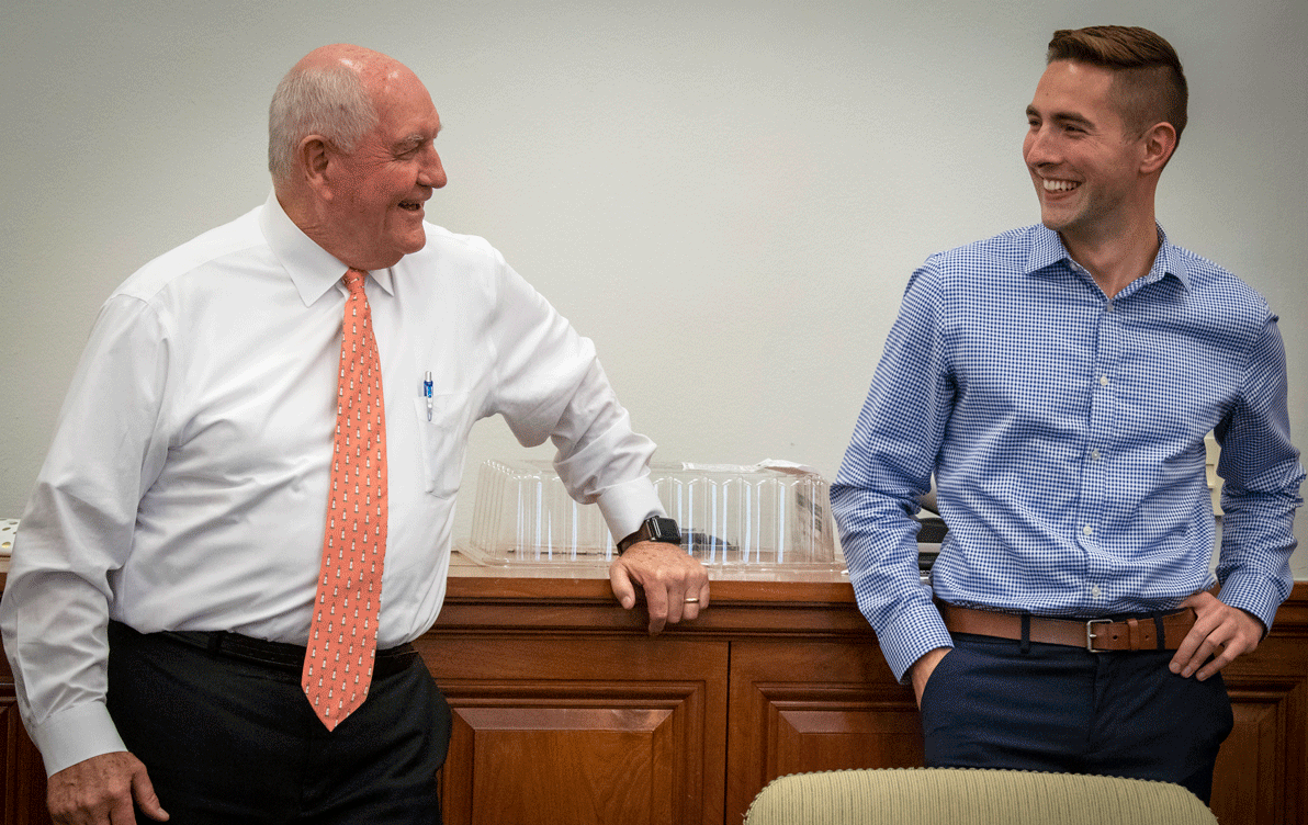 Two white men, one old wearing white shirt and orange tie, the other young wearing a blue shirt, stand in front of a wooden cabinet smiling at each other
