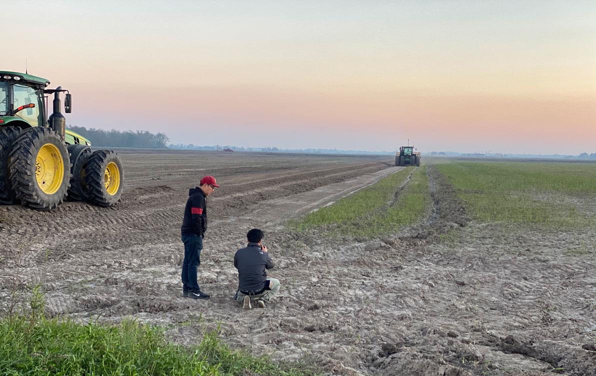 Tow men film combines on a barren field at sunset