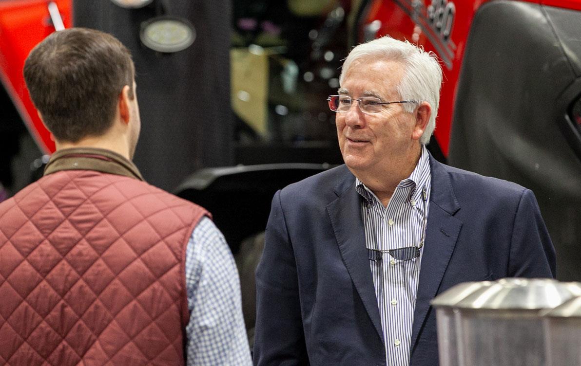 Older white man with white hair talks to younger man with his back to the camera