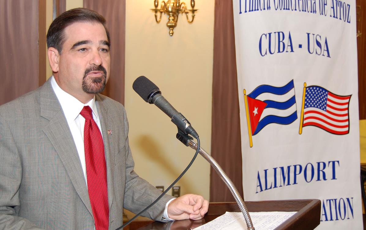White man wearing business suit & red tie stands at podium next to poster with US and Cuba flags