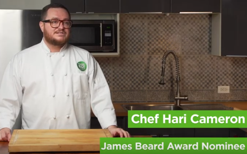 Chef Hari Cameron wearing white chef jacket with green Think Rice logo stands in kitchen, cutting board on table in front of him