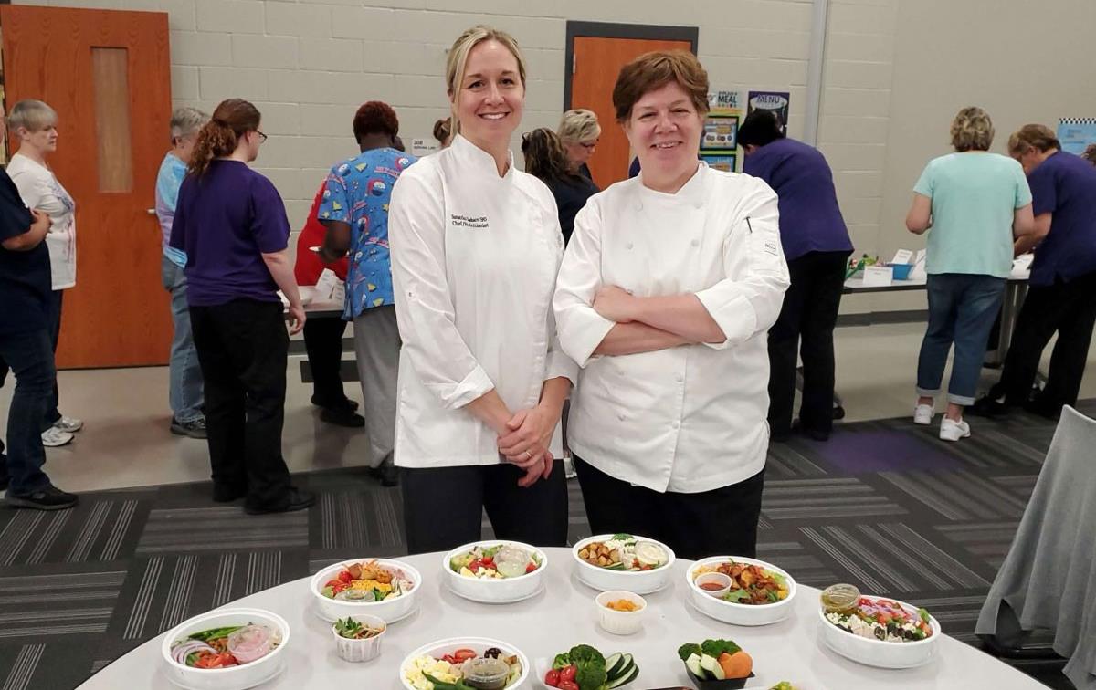 Two chefs in school cafeteria stand in front of table filled with an array of healthy student meals