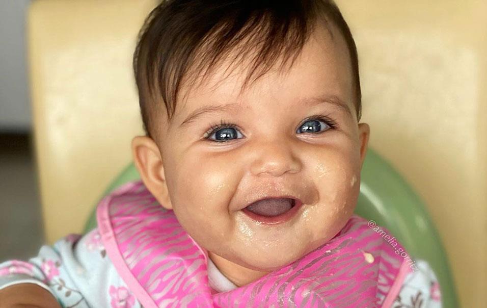 Smiling baby in high chair wearing pink bib with hand on package of infant rice cereal