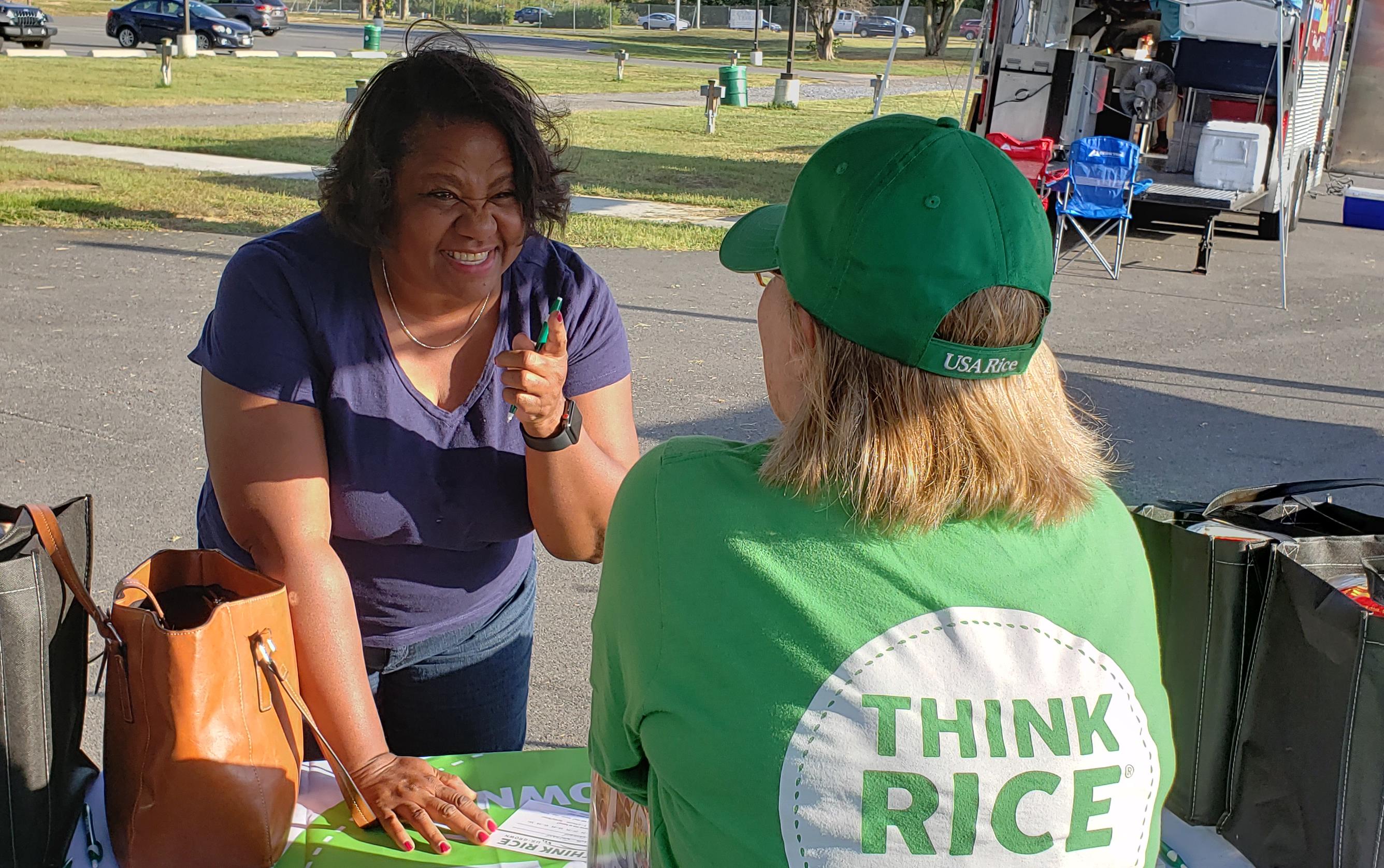 Woman stands across a table and shakes her finger at woman with back to camera wearing a green Think Rice shirt