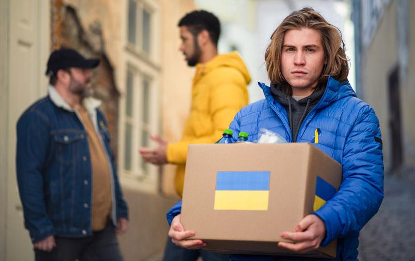 Young man carrying box of food aid with Ukrainian flag on each side of box, two men talking in background