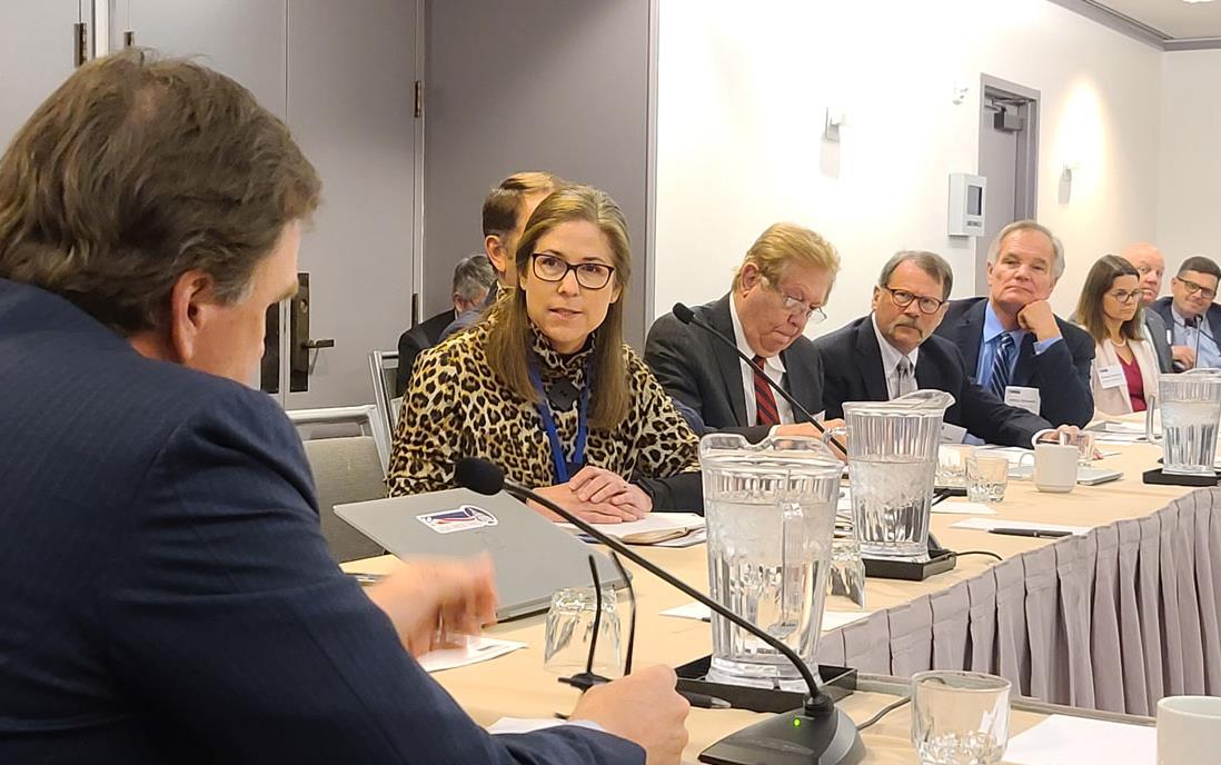 People sit at conference room table in front of microphones, pitchers of water, and laptops