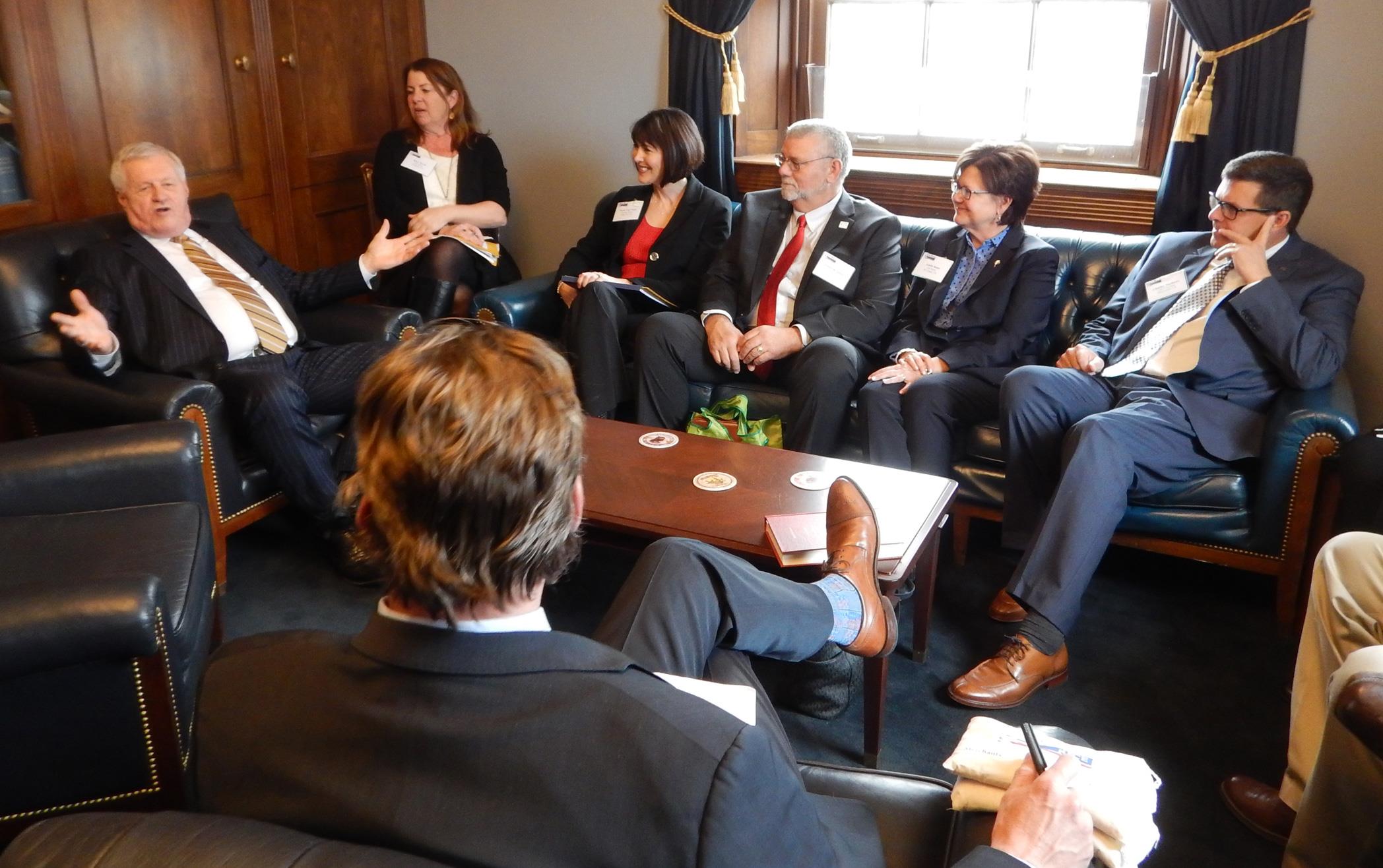 Group of people crowded into conference room, sitting on couches listening to white man in suit talk