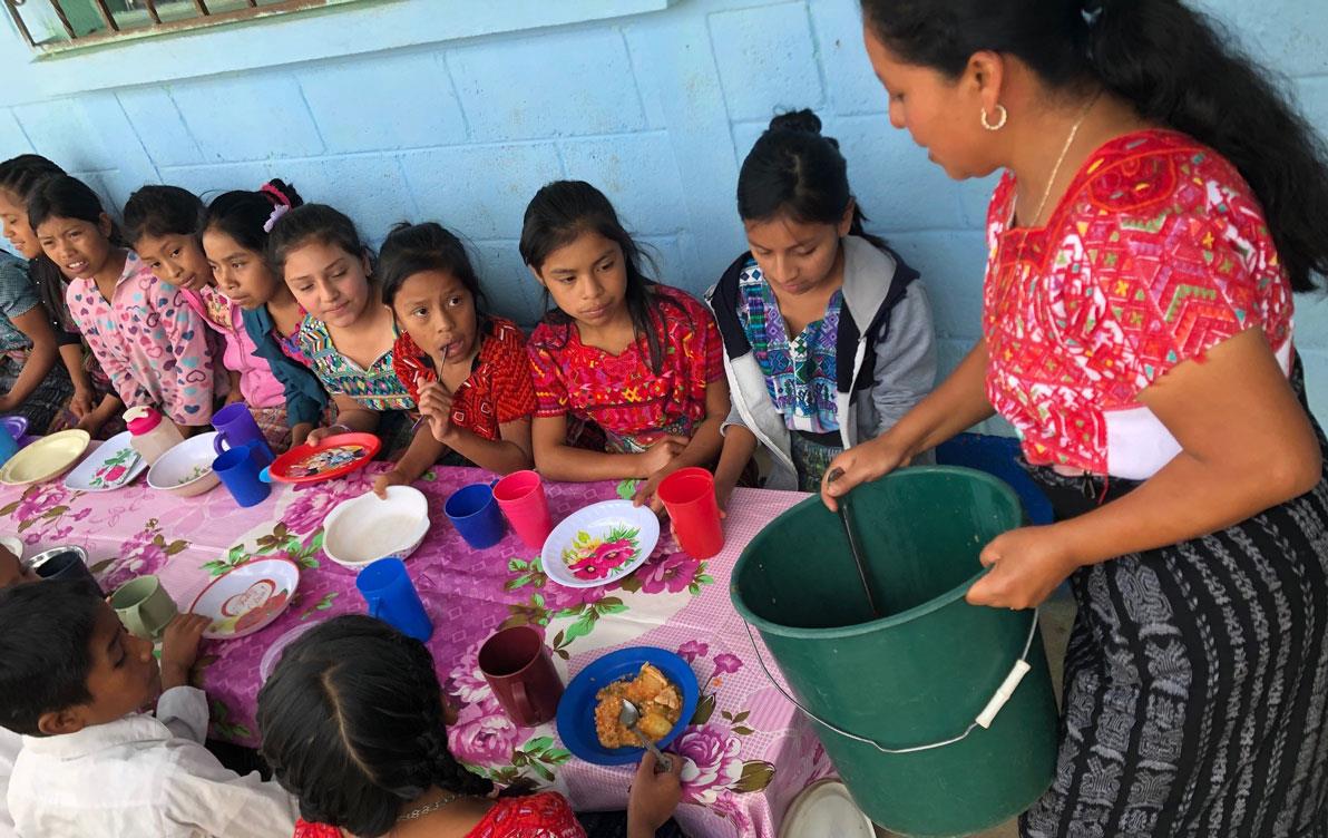 Children sit at a table, woman prepares to dish out food from a green bucket, everyone is colorfully dressed