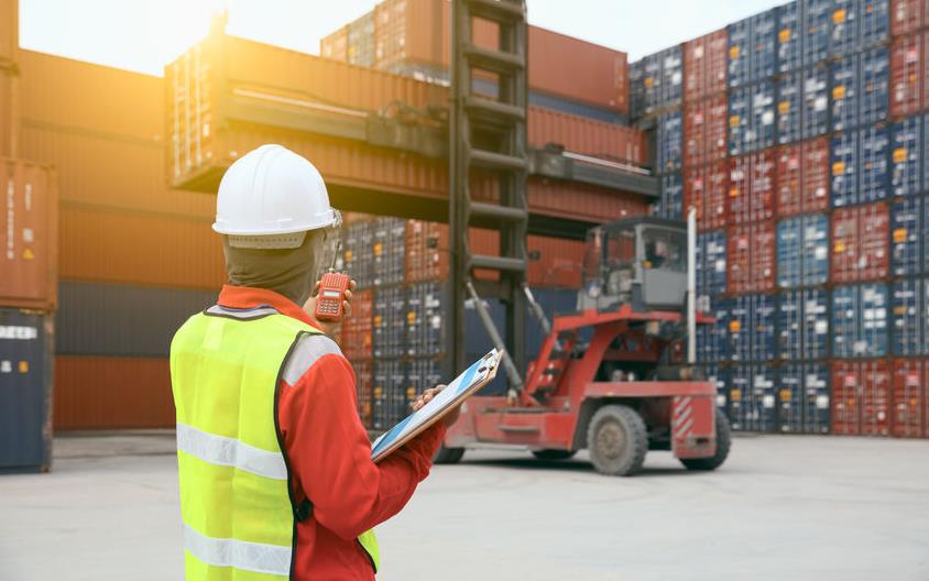 Foreman wearing safety vest, holding clipboard and cellphone, stands in front of forklift moving shipping containers