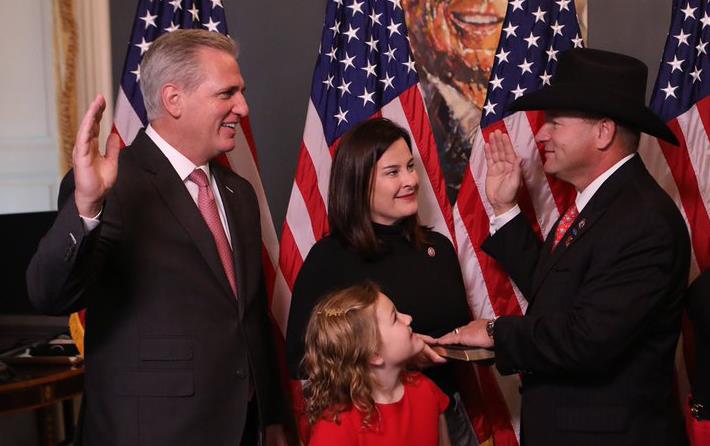 Rep Troy Nehls swearing ceremony shows two men in dark business suits, raising their right hands, surrounded by woman and three girls, everyone stands in front of American flags
