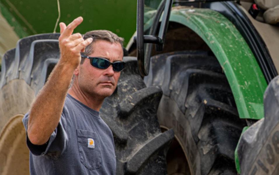 White man wearing gray t-shirt and sunglasses stands next to large combine tires