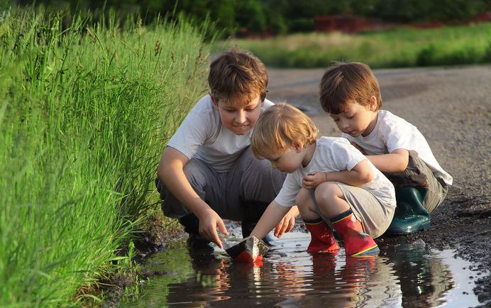 Children play with homemade sailboat in puddle next to overgrown, green, grassy area