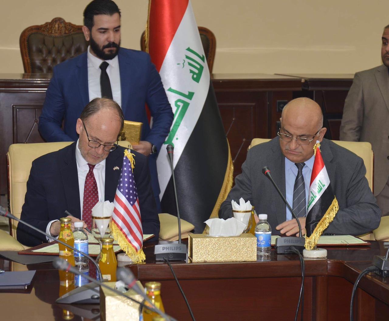 Two men sit at desk signing documents, US and Iraq flags in background