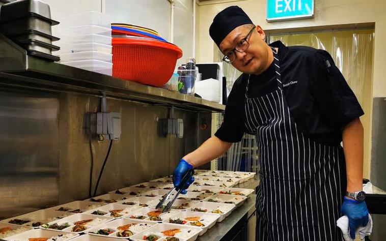 Chef wearing black apron with white stripes uses tongs to prep takeout meals in plastic containers