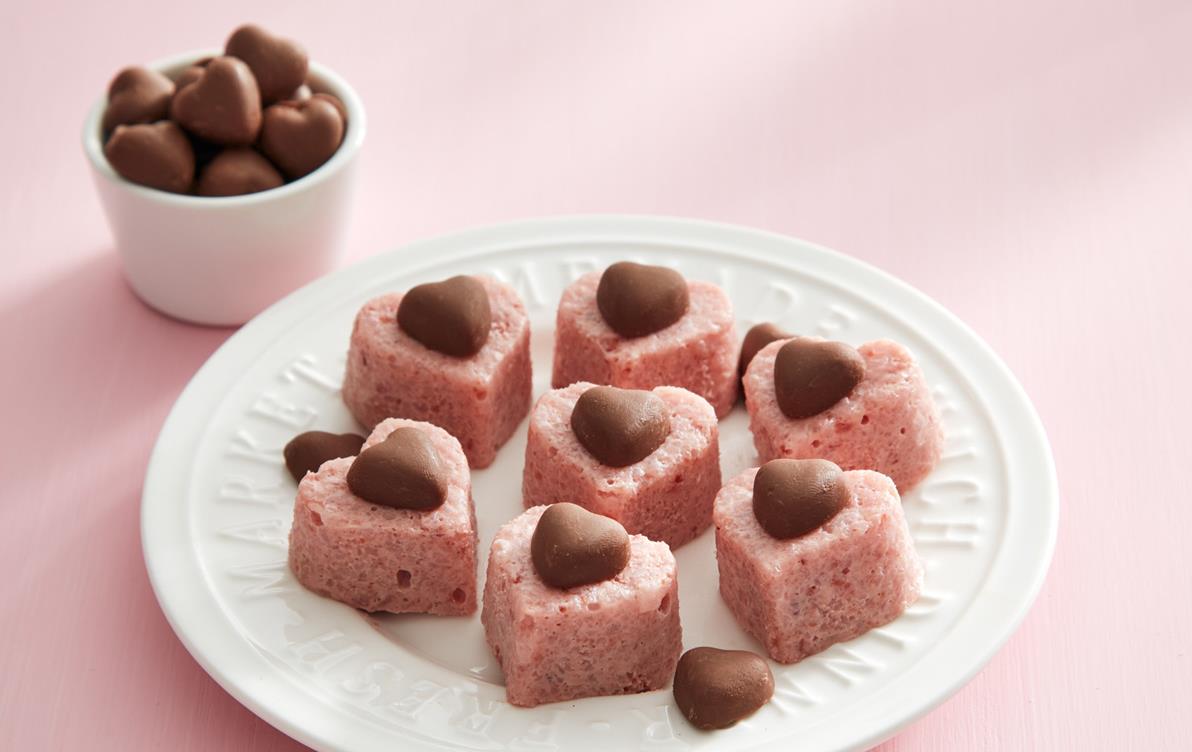 Pink heart-shaped rice croutons with chocolate hearts on top sit on white plate, a cup of chocolate hearts in background