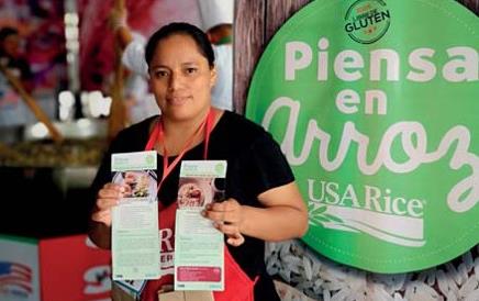 Guatemala promotion, woman wearing red apron holds brochures stands in front of Piensa en Arroz sign