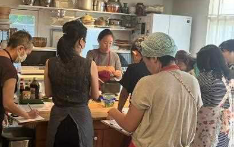 People wearing aprons and face masks gather around kitchen island to watch chef prepare rice dish