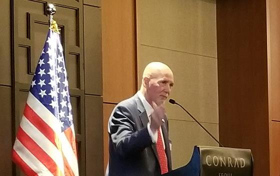 Korea Trade Mission, Ken Isley speaks from podium standing in front of US flag, seated officials at a table to his left