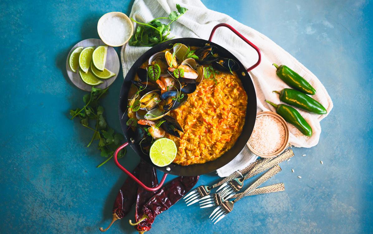 Overhead shot of Milpa-Paella with rice & seafood in black pan surrounded by silver forks, red & green chilis, and lime slices on blue background