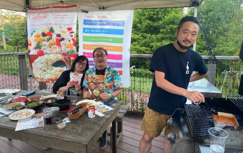 People sit at picnic table watching man standing at grill, colorful posters in background