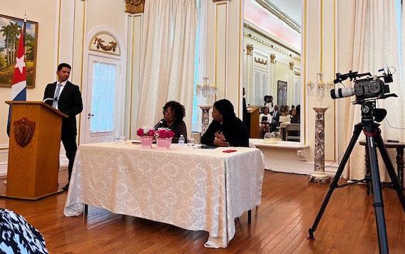 Poet-Nancy-Morejon-&-Cuban-Ambassador-Lianys-Torres-Rivera seated at table in ornate ballroom, cameraman w/set-up at right