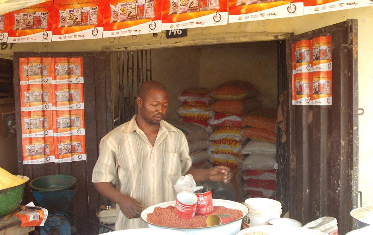 Open-air-market-in-Nigeria with USAR posters displayed on door frame where man stands behind table filled with large bowls of rice & beans