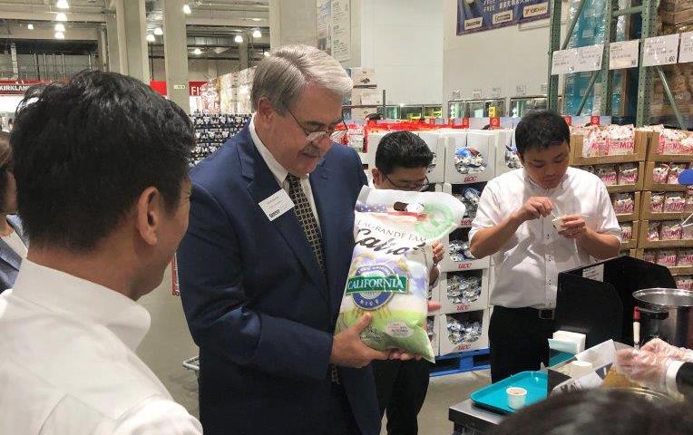 Man with gray hair wearing dark business suit holds large bag on rice while standing in grocery store aisle surrounded by group of people