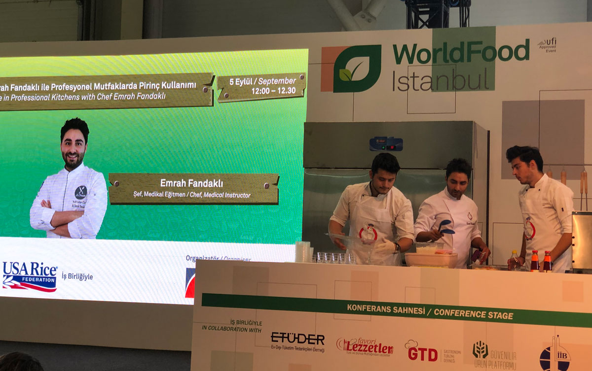 Three chefs cook on stage beside large photo of another chef with the USA Rice logo