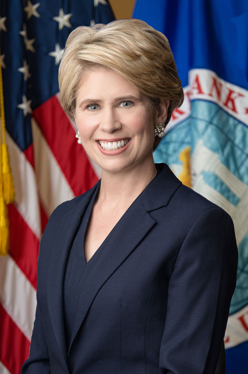 EXIM-Chair-Kimberly-Reed,-headshot, white woman with short blonde hair wearing dark business suit standing in front of US flag